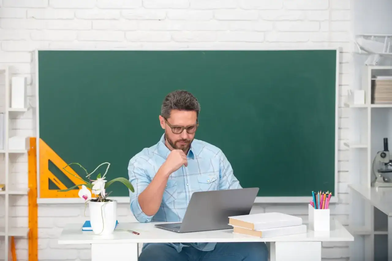 Nauczyciel przy biurku z laptopem, zastanawia się, czy uczniowie dostaną laptopy. Na biurku kwiat, książki i przybory.