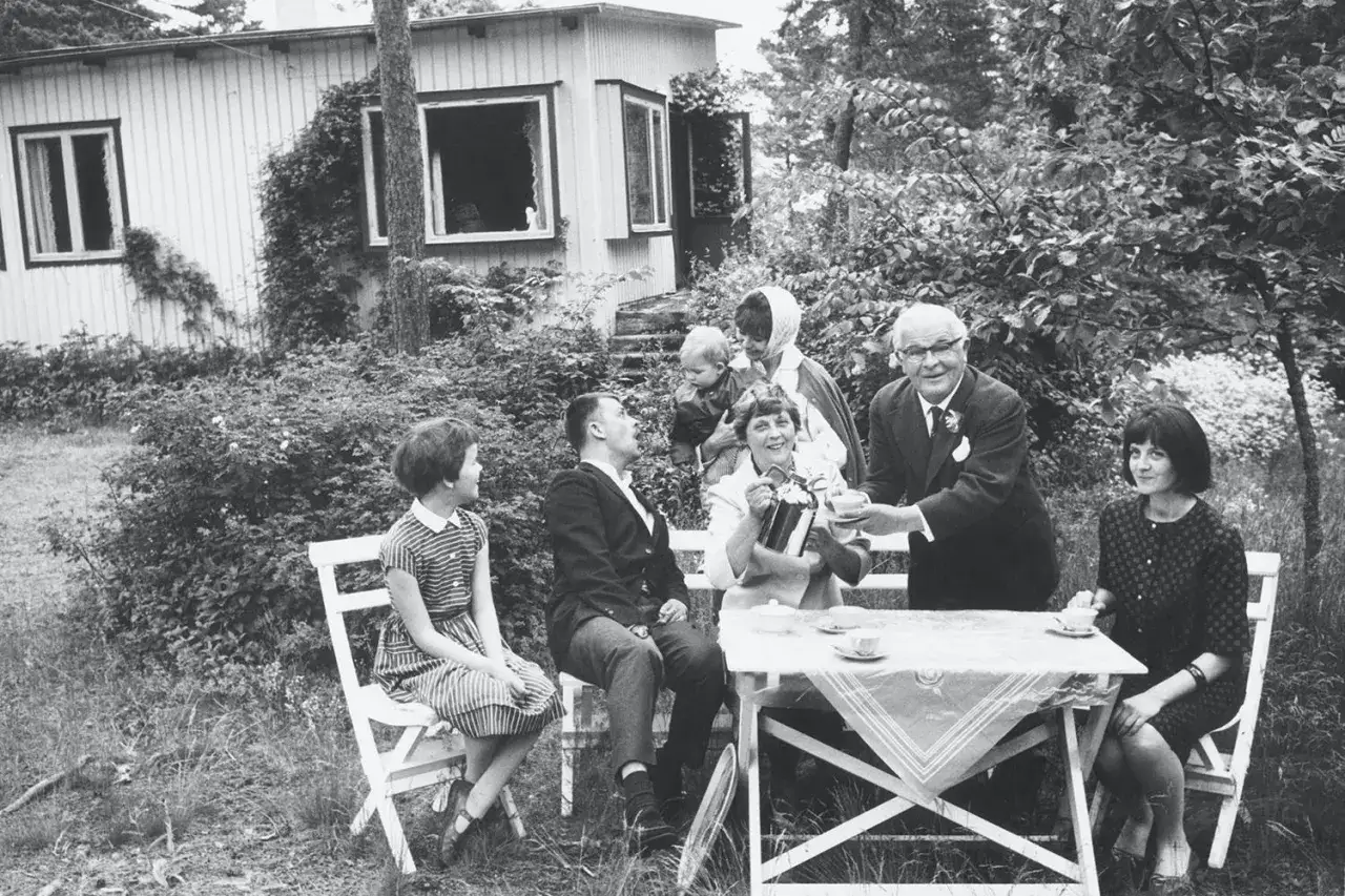En familj samlad för fika utomhus, kanske en festlig tillställning som påminner om en scen ur en Evert Taube-visa med barn och vuxna.