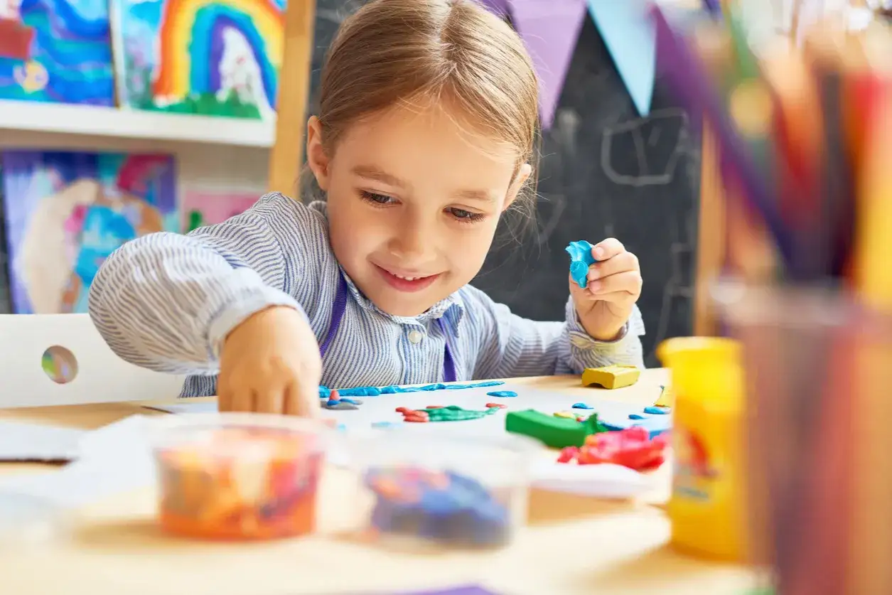 Ni&ntilde;a de 0-3 a&ntilde;os sonr&iacute;e mientras moldea plastilina azul. En la mesa hay m&aacute;s plastilina de colores y dibujos.