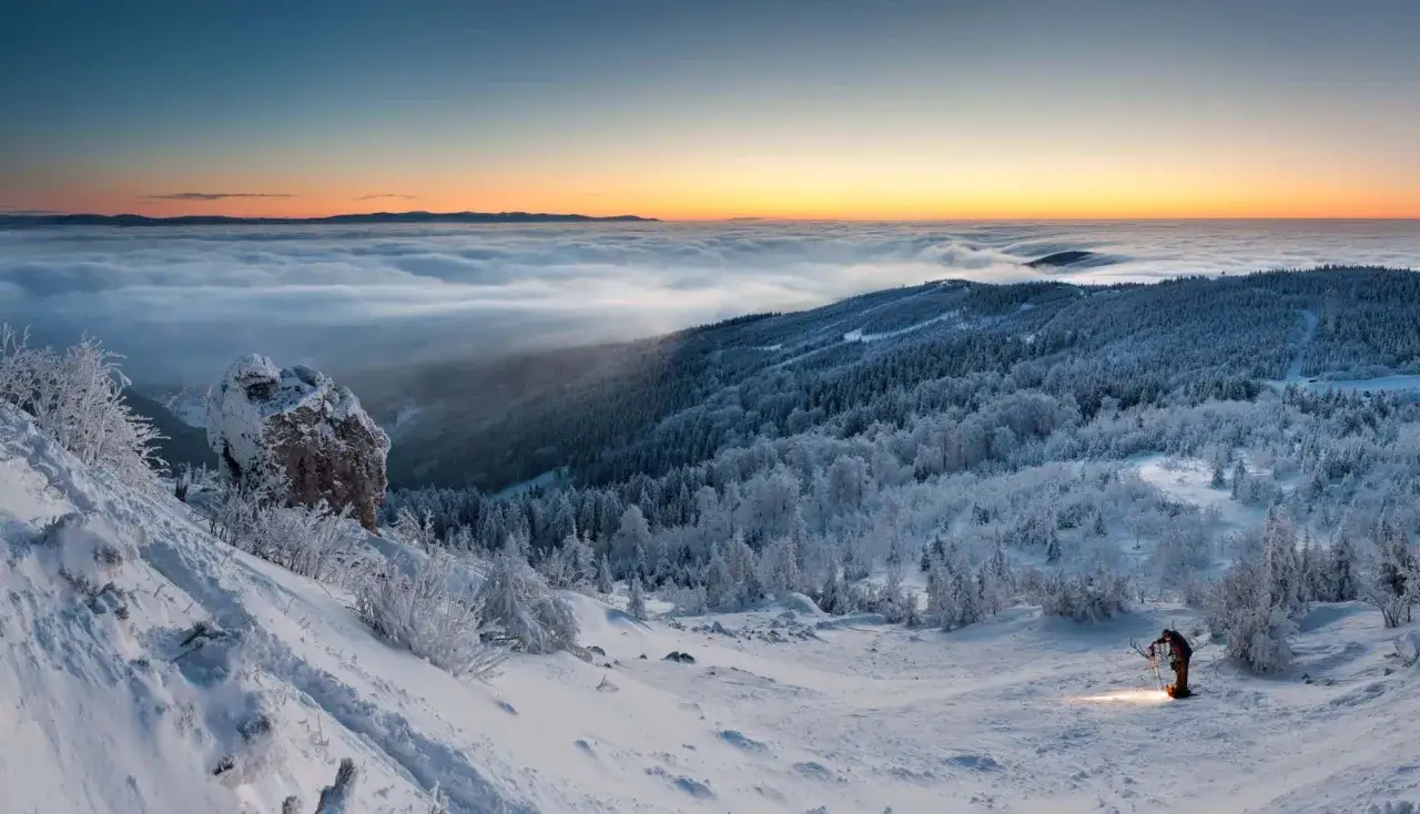 Ein einsamer Wanderer mit Stirnlampe genießt den atemberaubenden **Tschechien Winter**-Ausblick über einer Wolkendecke bei Sonnenaufgang.