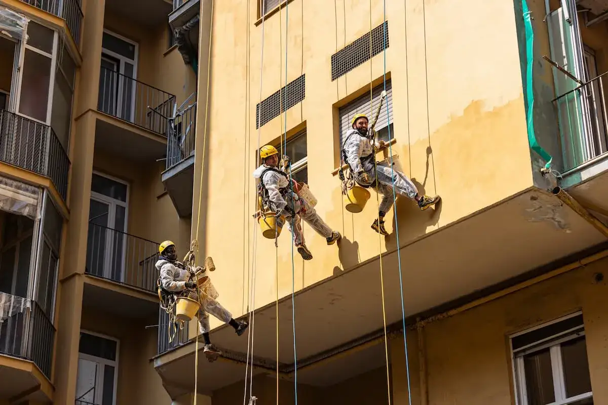 Operai in corda applicano il miglior intonaco per esterni su un edificio giallo. Lavorano in sicurezza, con imbracature e secchi.