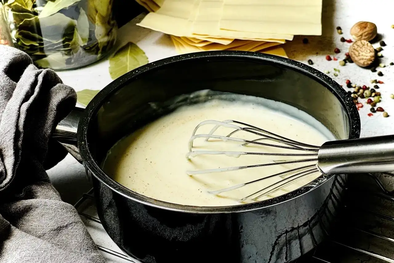 Cremige b&eacute;chamelsauce mit Sahne wird in einem Topf mit einem Schneebesen zubereitet. Daneben liegen Lasagneplatten und Gew&uuml;rze.