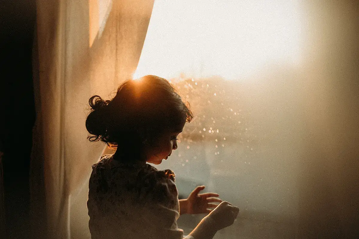 Bambina con capelli raccolti, illuminata dal sole che filtra attraverso una tenda. Gioca con la luce e ombra immagini, creando un'atmosfera magica.