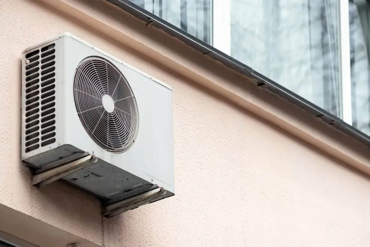 Biała klimatyzacja na jasnoróżowej elewacji budynku, zamontowana na wspornikach. Widać kratkę wentylacyjną i okrągły wentylator.