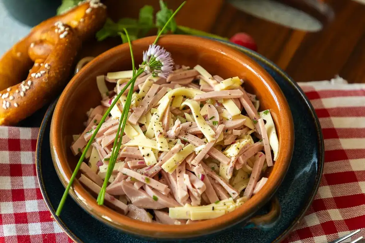 Schmackhafter Schweizer Wurstsalat mit Käse, Zwiebeln und Kräutern, garniert mit einer Schnittlauchblüte. Dazu ein Laugenbrezel.
