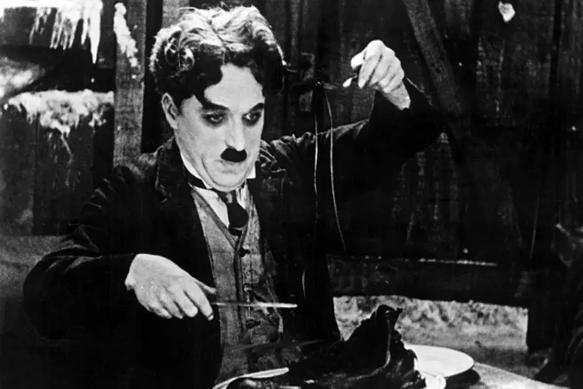 Charlie Chaplin, en ikonisk figur från en chaplin film klassiker, sitter vid ett bord och undersöker en sko med en kniv.