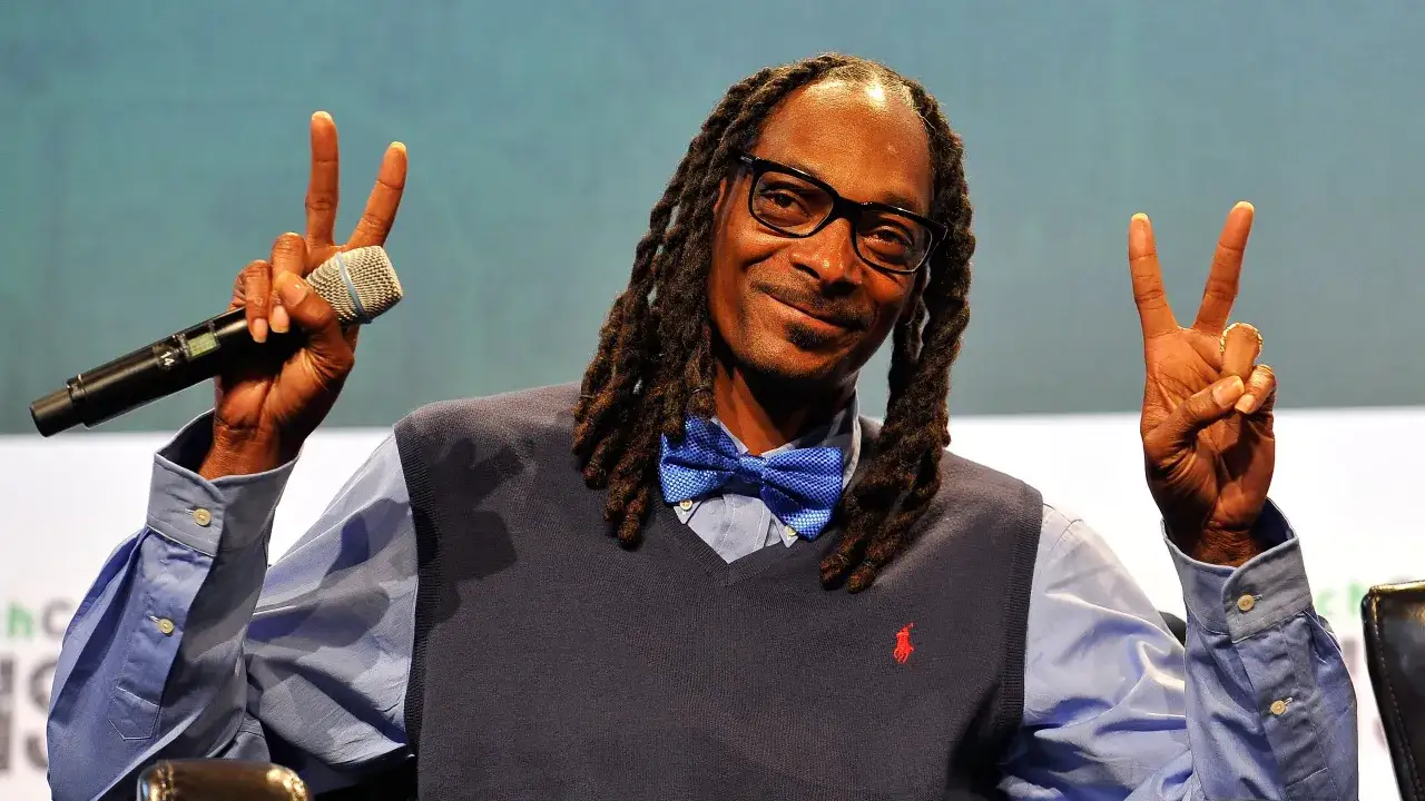 Snoop Dogg z mikrofonem i dwoma palcami w g&oacute;rze, jakby pytał 