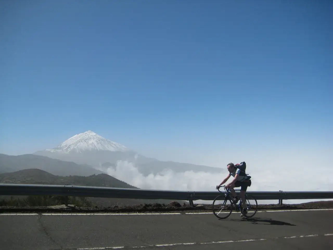 Ein Radfahrer auf Teneriffa mit Rennrad und Rucksack f&auml;hrt auf einer Stra&szlig;e. Im Hintergrund thront der schneebedeckte Teide.