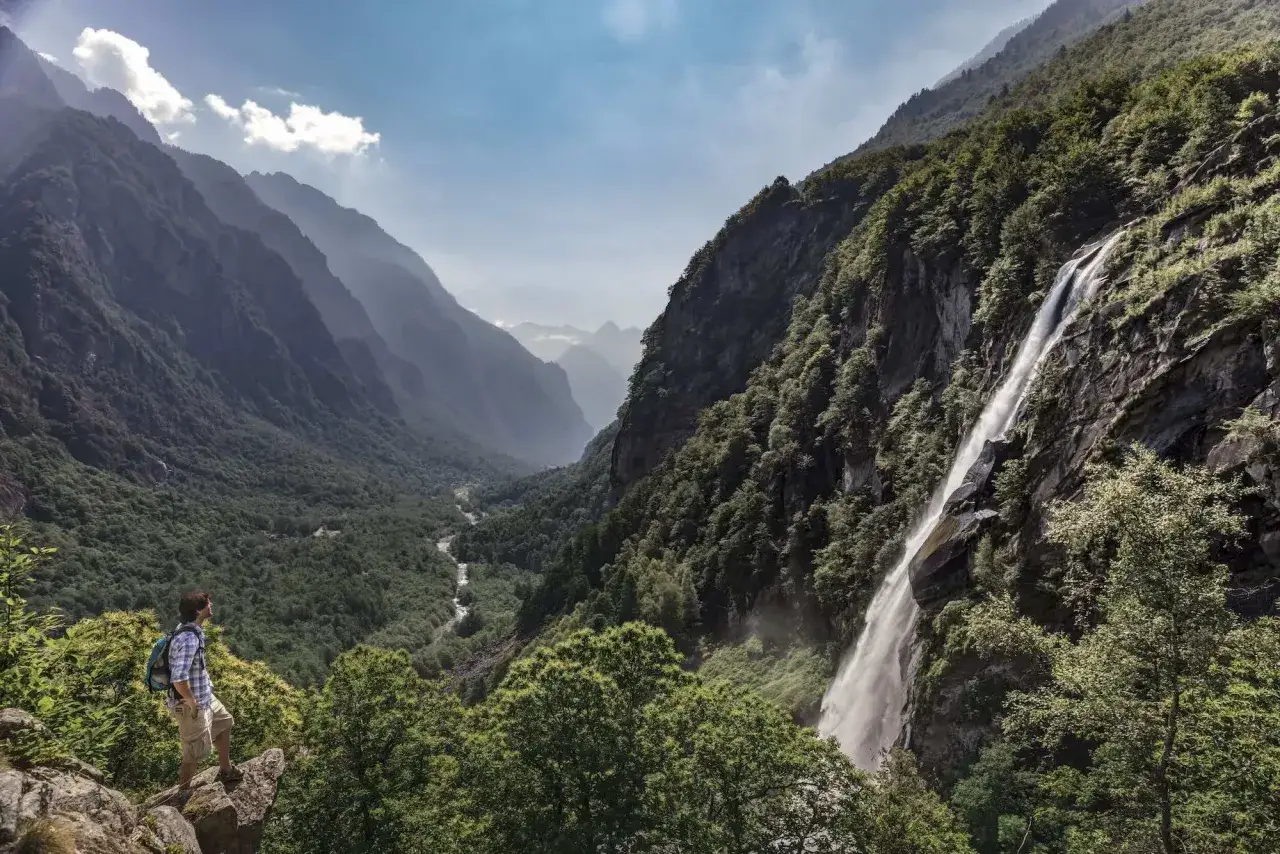 Ein Wanderer genie&szlig;t die Aussicht auf einen Wasserfall und ein gr&uuml;nes Tal im Tessin. Perfekt f&uuml;r Tessin Wandern.