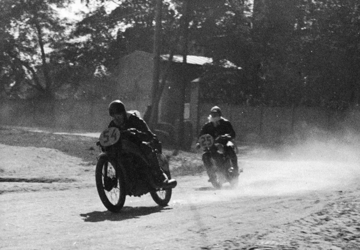 Dwóch motocyklistów ściga się po piaszczystej drodze, unosząc tumany kurzu. To fragment historii motocykla, pełen adrenaliny i ducha rywalizacji.