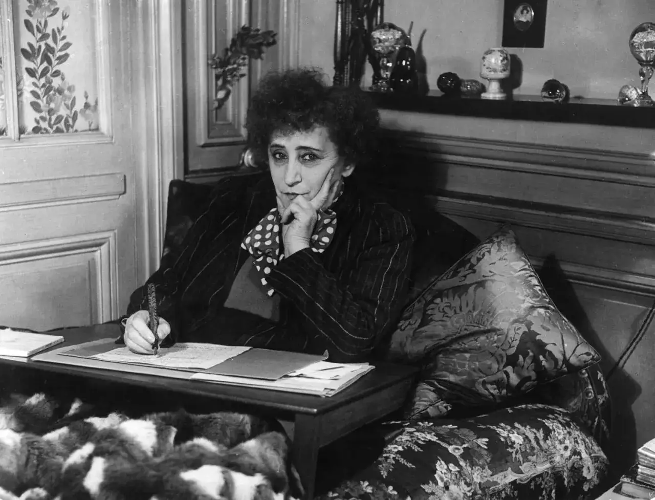 Colette, z burzą włosów i w kropkowanej chuście, pisze przy biurku. W jej oczach widać skupienie, a dłoń z piórem spoczywa na papierze.
