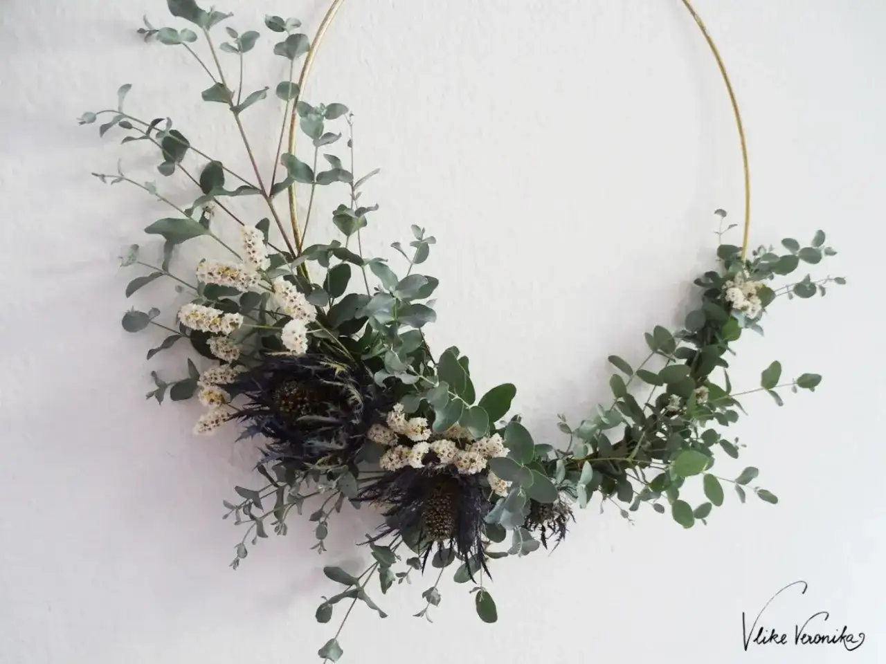 Ein moderner Blumenkranz mit Eukalyptus, kleinen weißen Blüten und dunklen Disteln. Ein schönes Beispiel, wie man einen Blumenkranz binden kann.