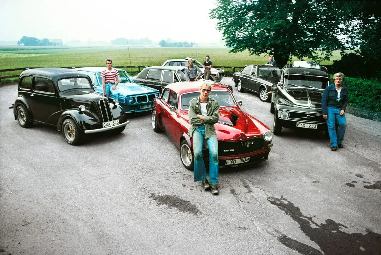 En grupp unga män poserar med sina klassiska sportbilar. En röd Volvo P1800, en blå Volvo Amazon och en svart Ford Anglia står uppradade.