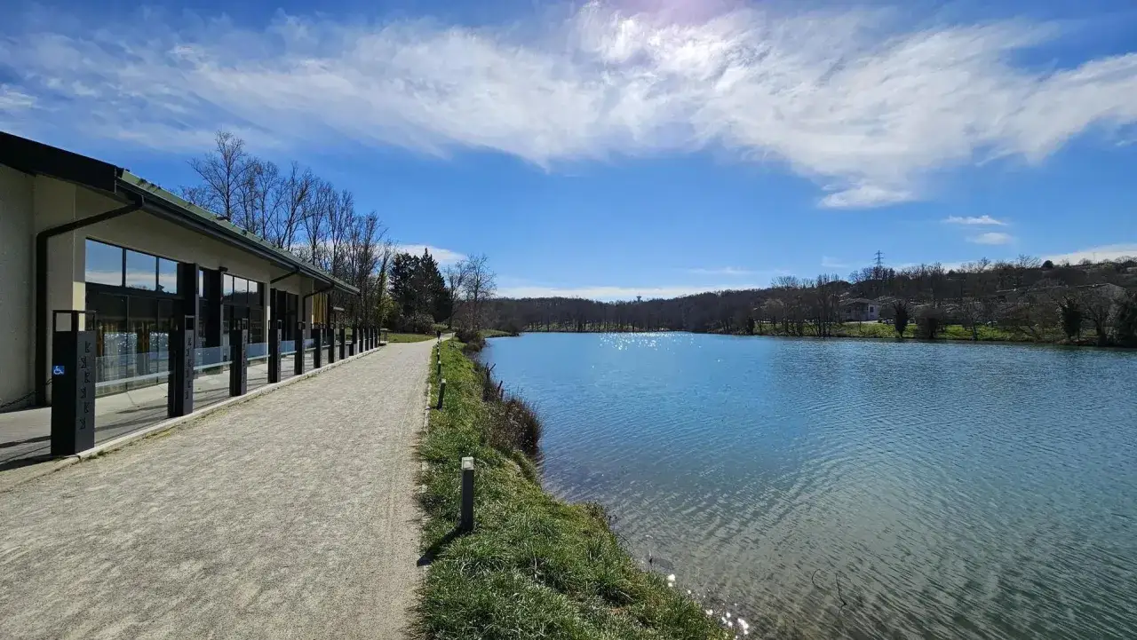 Le lac de Flourens, bordé par un chemin et un bâtiment moderne, reflète un ciel bleu éclatant sous un soleil radieux.