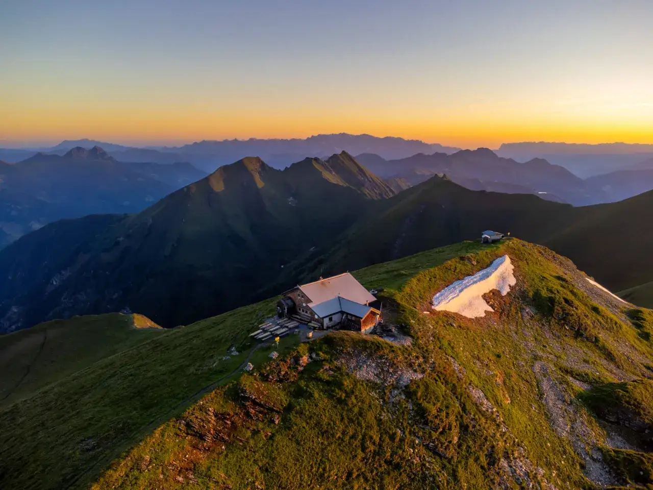 Sonnenuntergang &uuml;ber den Alpen. Eine Bergh&uuml;tte, ideal f&uuml;r mehrtagestouren, thront auf einem Gipfel, umgeben von sanften H&uuml;geln und dramatischen Bergketten.