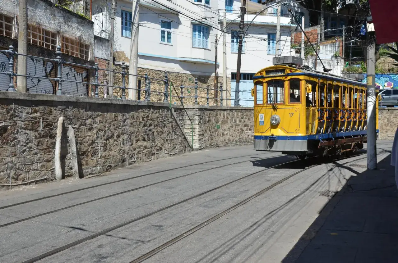 Gelber Stra&szlig;enbahnwagen f&auml;hrt durch eine Stra&szlig;e in Rio de Janeiro, vorbei an einer Steinmauer und bunten H&auml;usern.