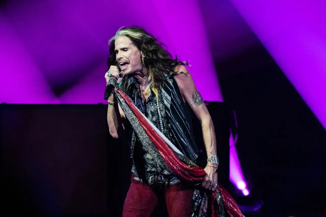 Steven Tyler, wokalista zespołu Aerosmith, śpiewa z pasją na tle fioletowych świateł scenicznych.