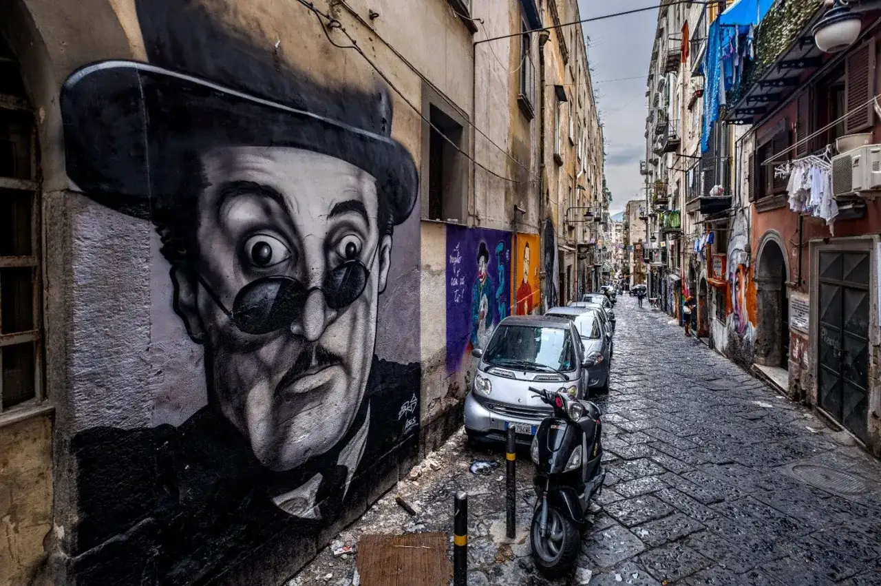 Un imponente murales napoli di un uomo con cappello e occhiali da sole domina un vicolo stretto, fiancheggiato da auto e scooter.