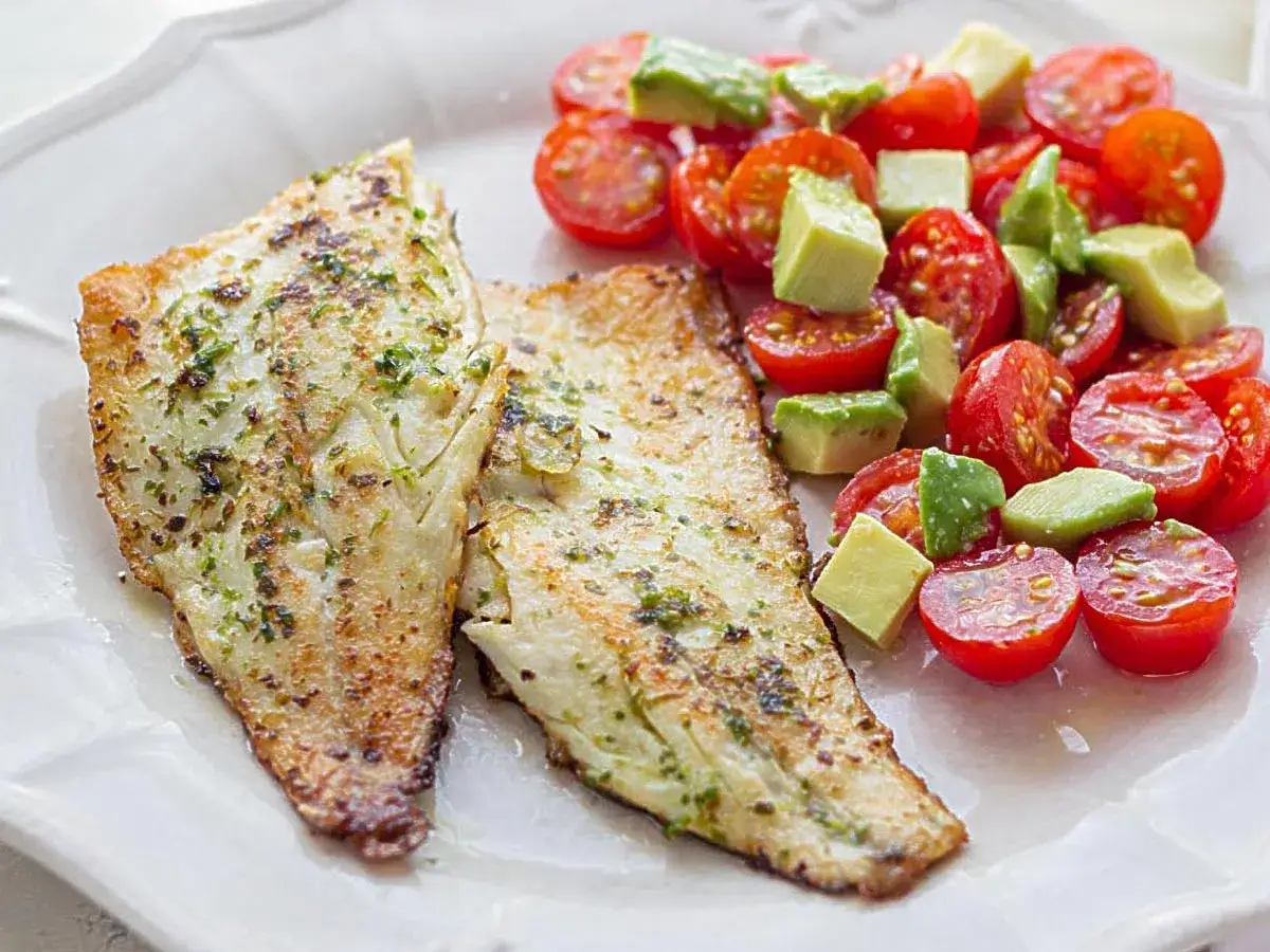 Dos filetes de lubina a la plancha con hierbas, acompañados de una ensalada fresca de tomates cherry y aguacate en cubos.