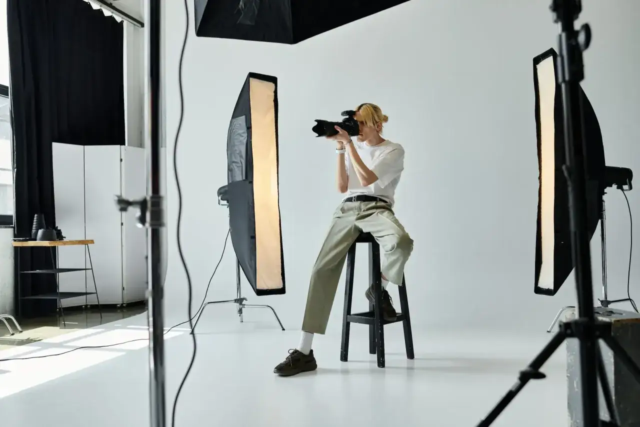 Młody fotograf z blond włosami siedzi na stołku, robiąc zdjęcie aparatem. Wokół niego studyjne oświetlenie. Zastanawiasz się, ile zarabia fotograf?
