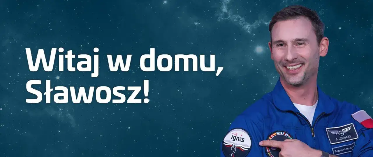 Uśmiechnięty Sławosz, polak w kombinezonie astronauty, wskazuje na naszywkę. Napis