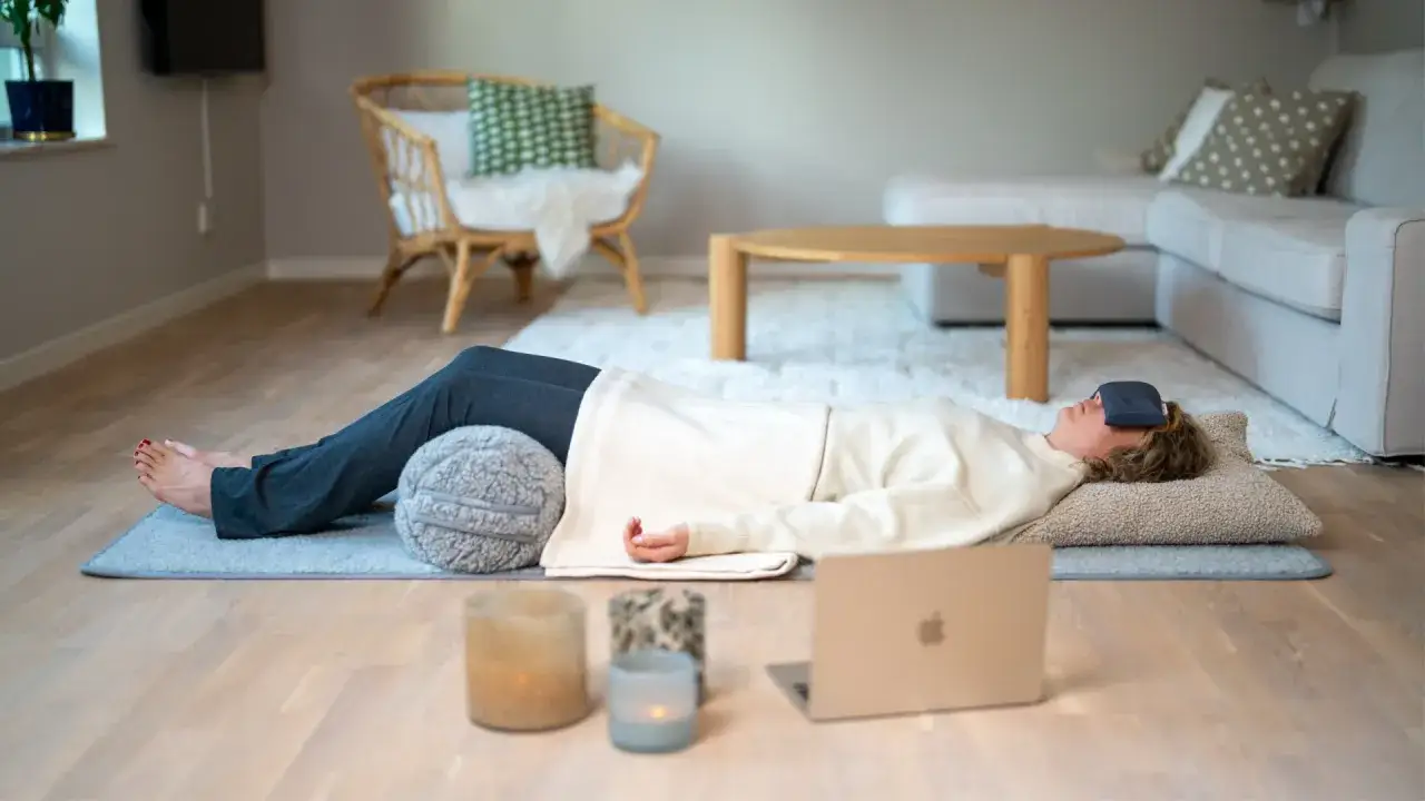 Person ligger på en matta med ögonbindel för att minska stress. Ljus och laptop i närheten.