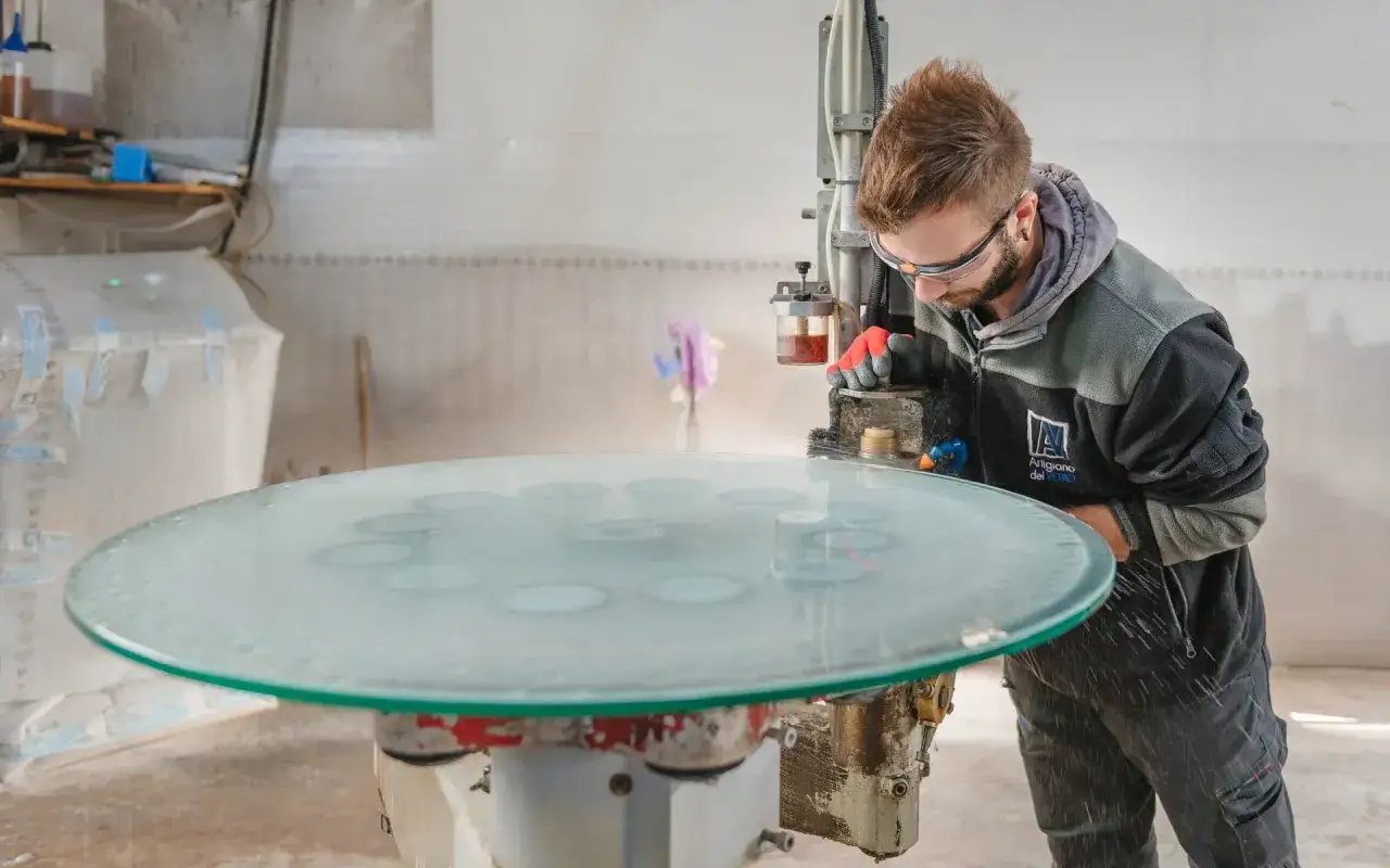 Artigiano al lavoro per molare il vetro di un grande piano circolare, con schizzi d'acqua e polvere di vetro.