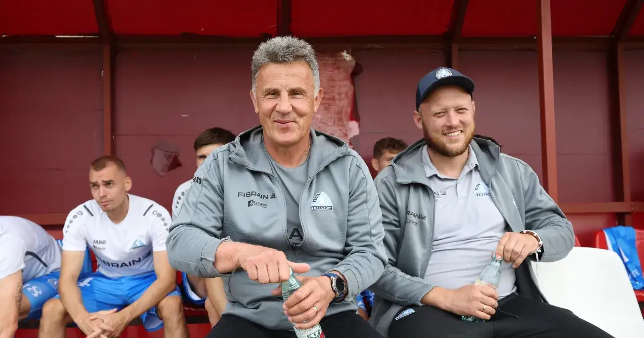 Marek Zub, trener Stali Rzeszów, z uśmiechem na twarzy, siedzi na ławce z butelką wody.