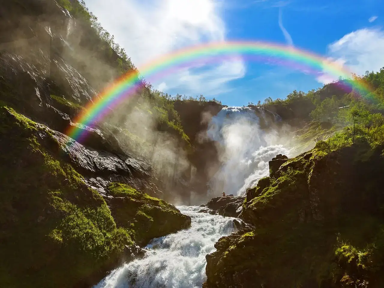 Majestätischer norwegischer Wasserfall, umrahmt von einem Regenbogen. Ein kleiner Mensch steht am Felsen und bestaunt die Naturgewalt.