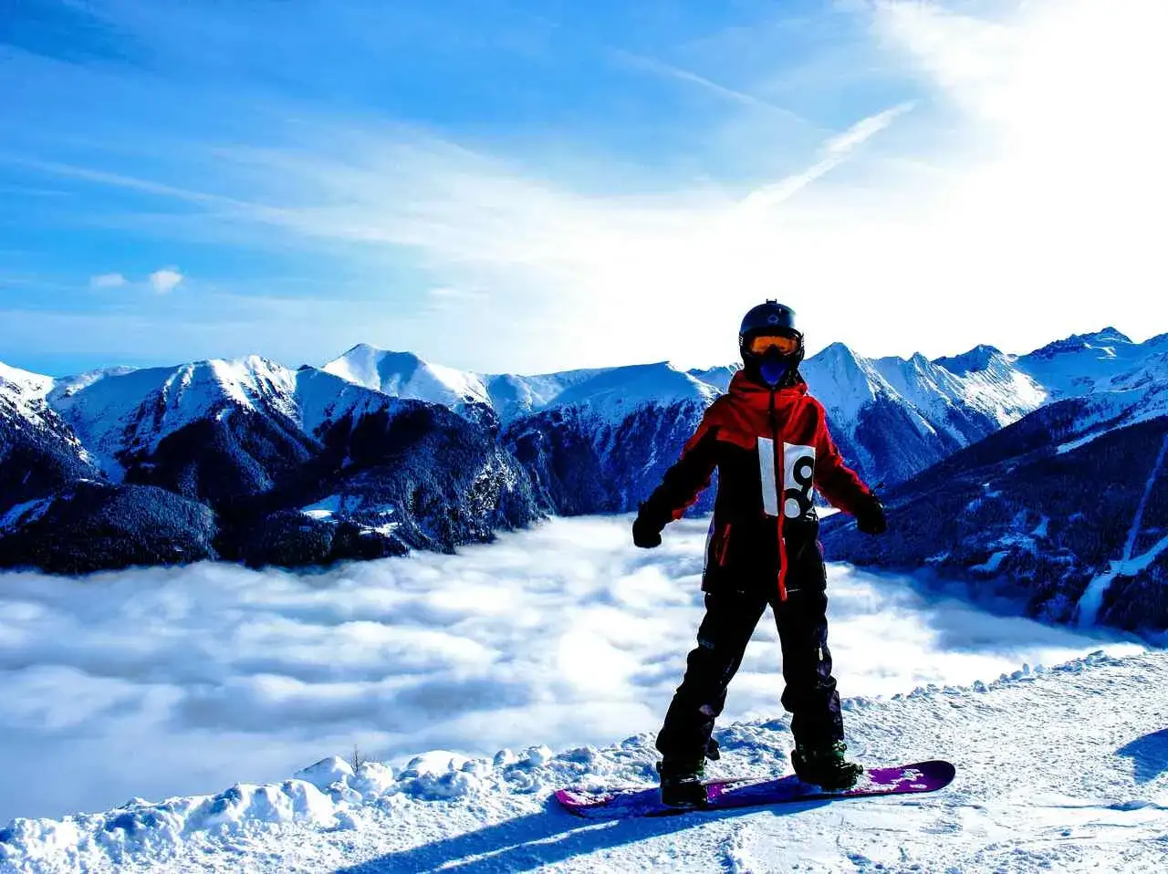Snowboardzista w czerwonej kurtce, która wygląda jak hardshell, stoi na szczycie góry. Poniżej morze chmur, a w tle ośnieżone szczyty.