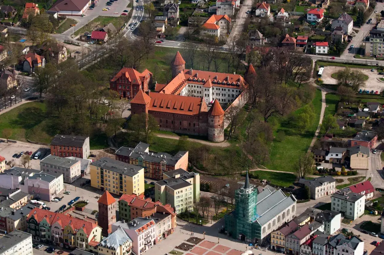 Zamek w Bytowie, potężna ceglana twierdza z czerwonymi dachami, otoczona zielenią i zabudowaniami miejskimi.