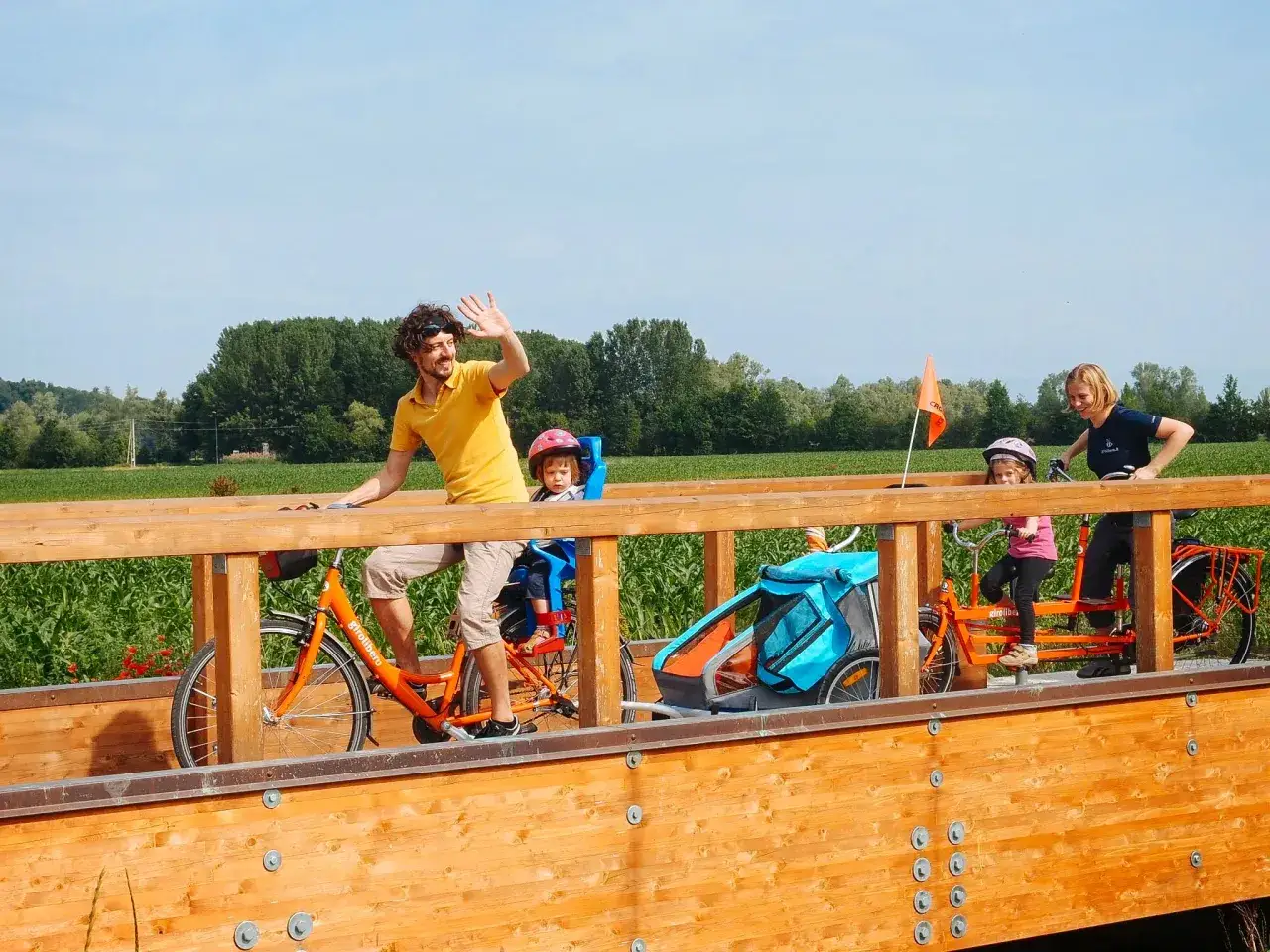 Famiglia in vacanze in bici con bambini su un ponte di legno, con un rimorchio per bambini e un tandem.