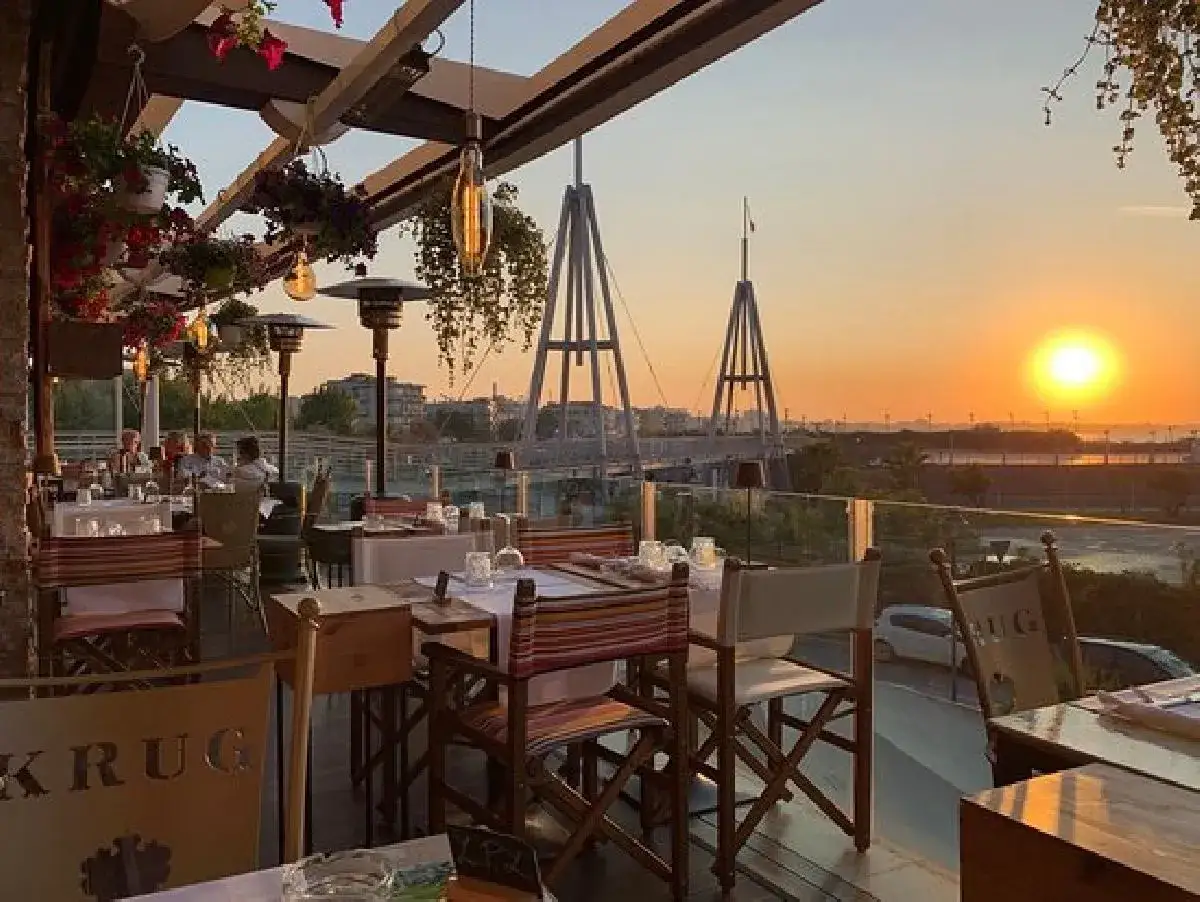 Tavoli apparecchiati in un ristorante a Rimini, con vista sul tramonto e ponti sullo sfondo. Atmosfera romantica per cene indimenticabili.