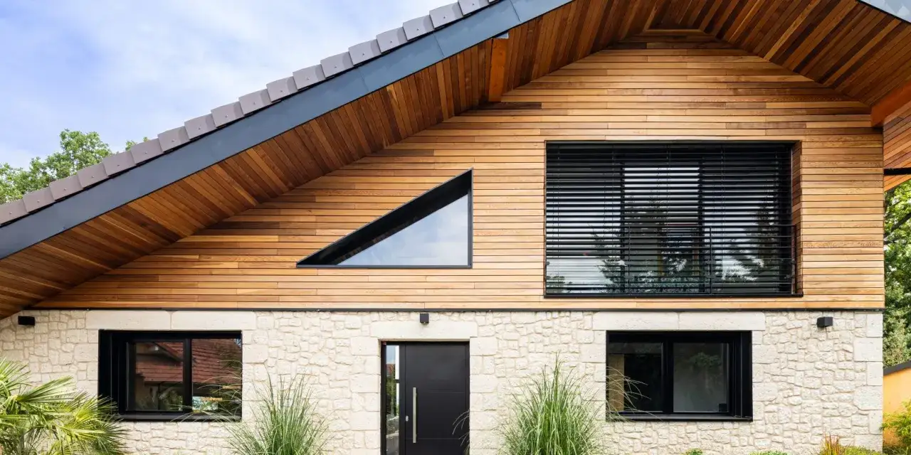 Ce type de maison moderne combine pierre claire et bois, avec une fen&ecirc;tre triangulaire audacieuse et de grandes baies vitr&eacute;es.