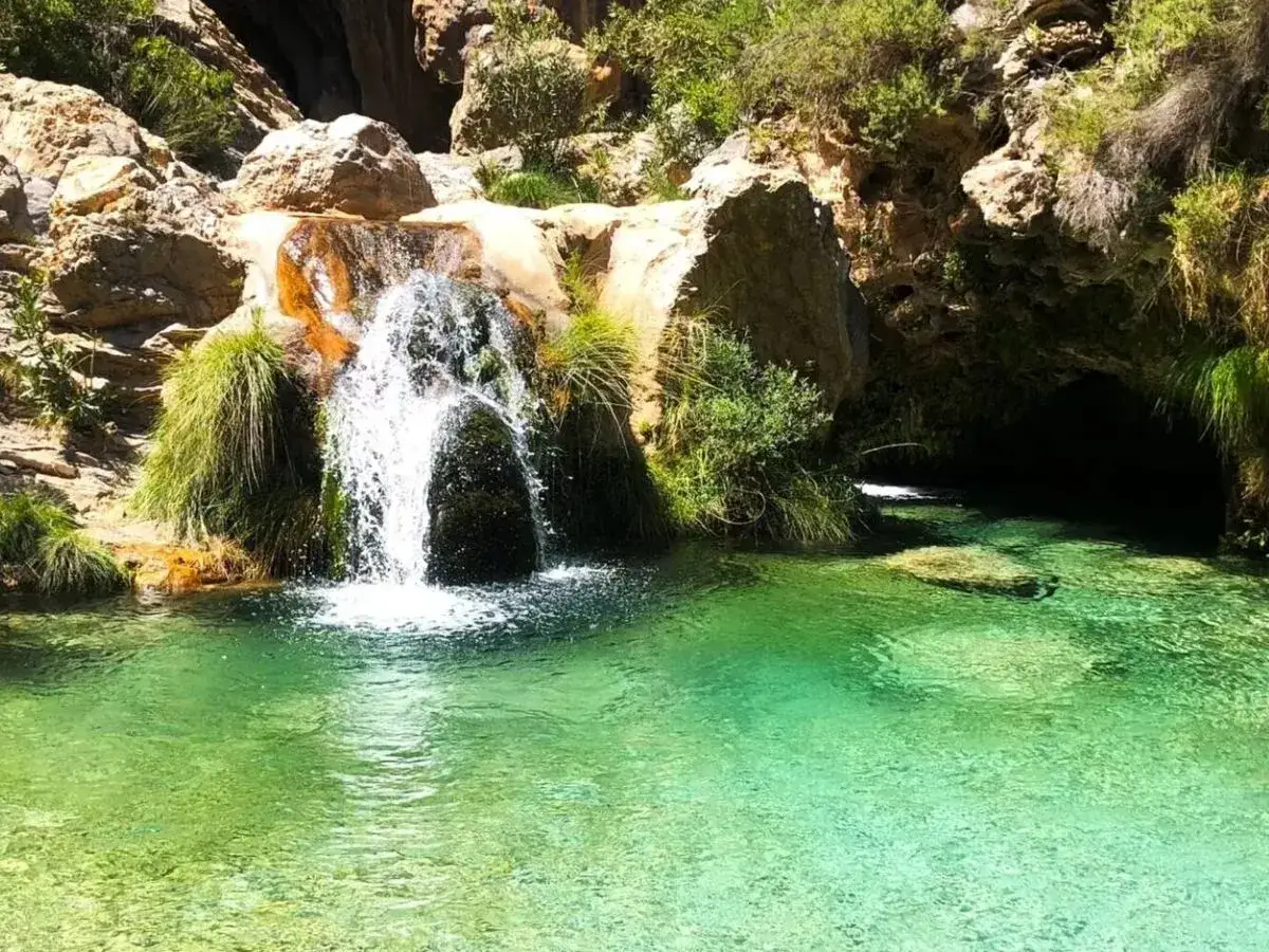 Cascada cayendo en unas pozas de agua cristalina y verde, rodeadas de rocas y vegetaci&oacute;n.