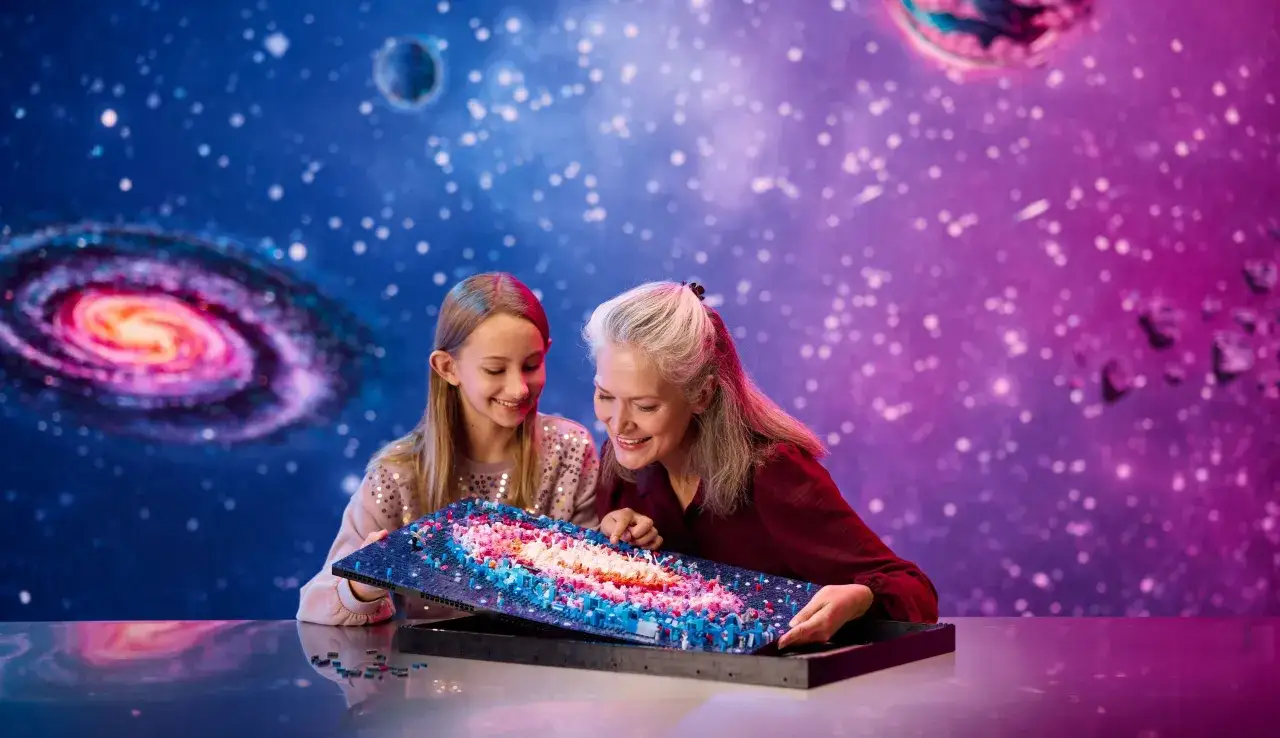 Zwei Frauen bestaunen ein detailreiches Milky Way Lego-Modell vor einem funkelnden Sternenhintergrund.