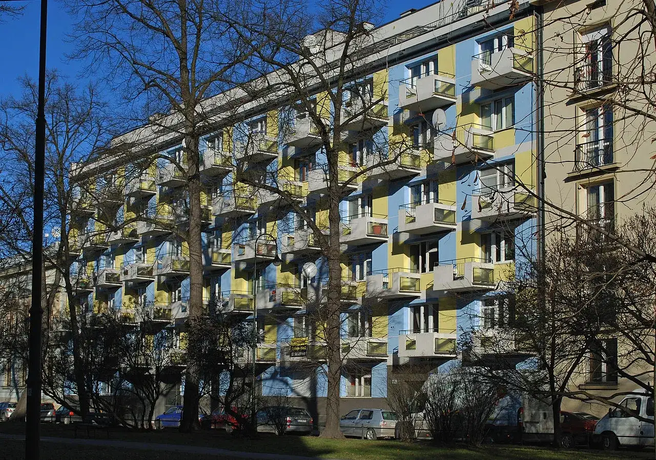 Charakterystyczna architektura PRL: wielopiętrowy blok mieszkalny z żółto-niebieskimi pasami i licznymi balkonami.