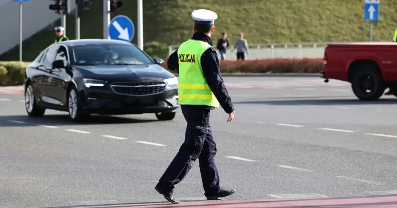 Policjant w kamizelce odblaskowej kieruje ruchem. Czarny samochód czeka, a obok widać czerwoną ciężarówkę. Dodatkowa masa w samochodzie nie jest widoczna.