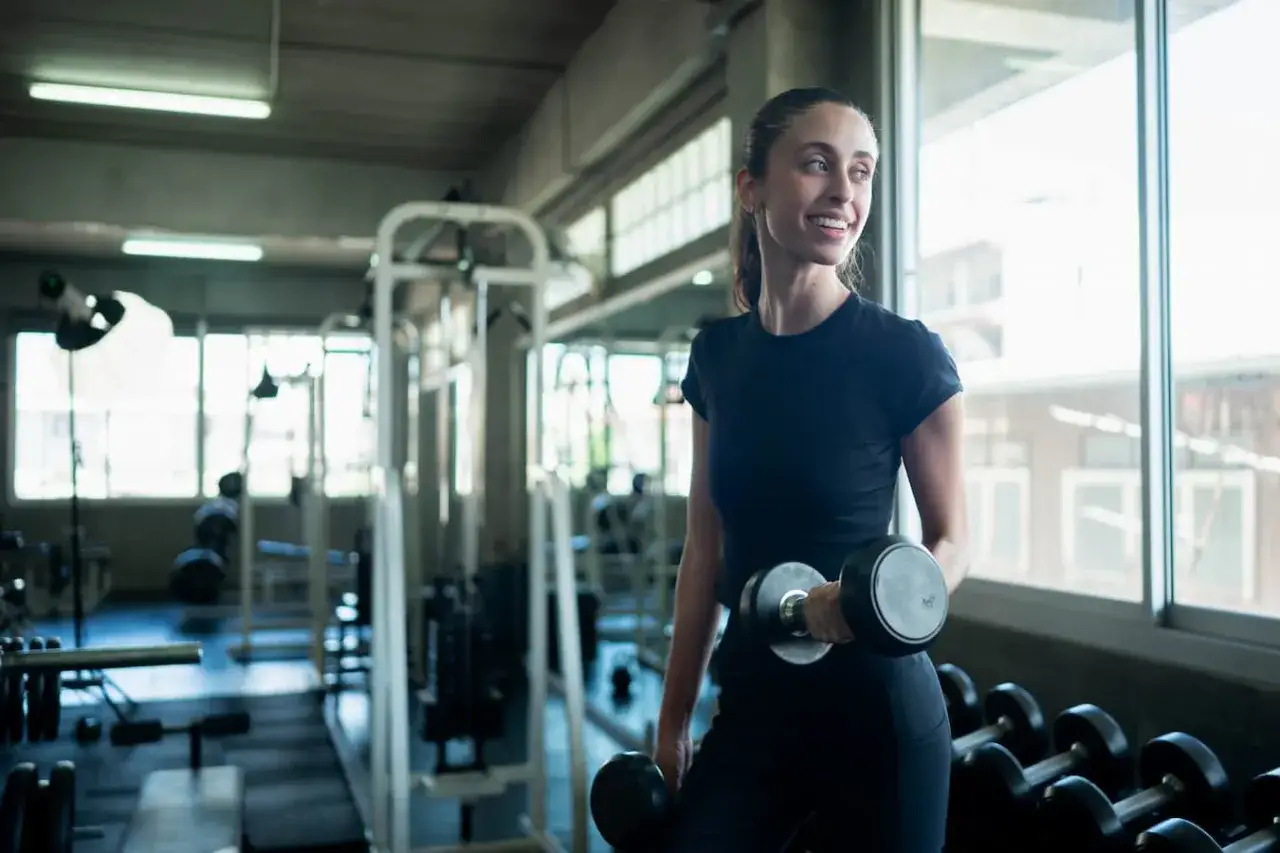 Donna sorridente in palestra solleva manubri, un esempio di come funziona il deficit calorico per raggiungere i propri obiettivi di fitness.