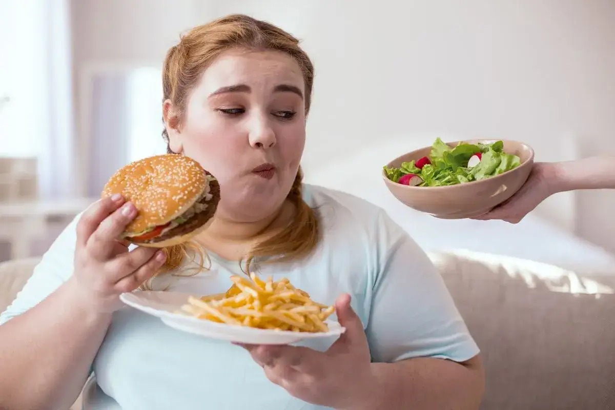 Kobieta z wahaniem patrzy na hamburgera i frytki, podczas gdy ktoś podaje jej miskę sałatki. To chyba odpowiedź na pytanie: dlaczego nie mogę schudnąć.