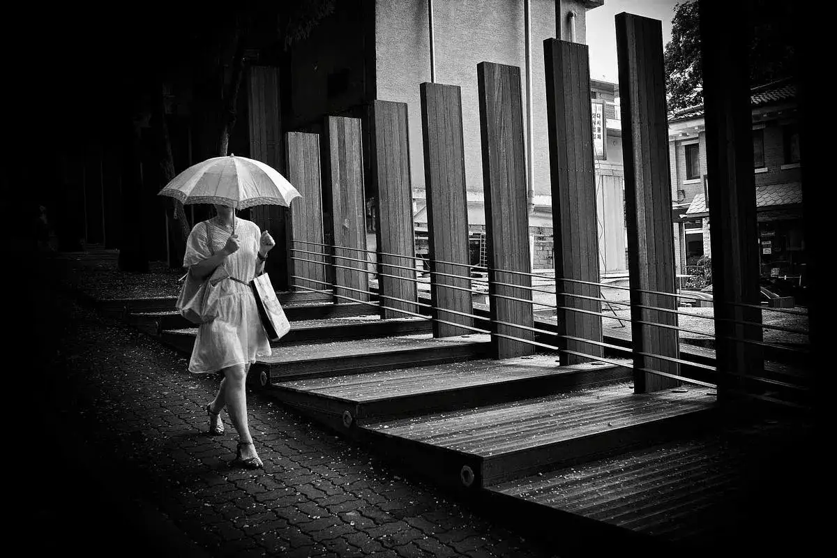 Kobieta z parasolką idzie po mokrym chodniku, tworząc dynamiczne rodzaje kompozycji w fotografii. Drewniane słupy i schody dodają głębi.
