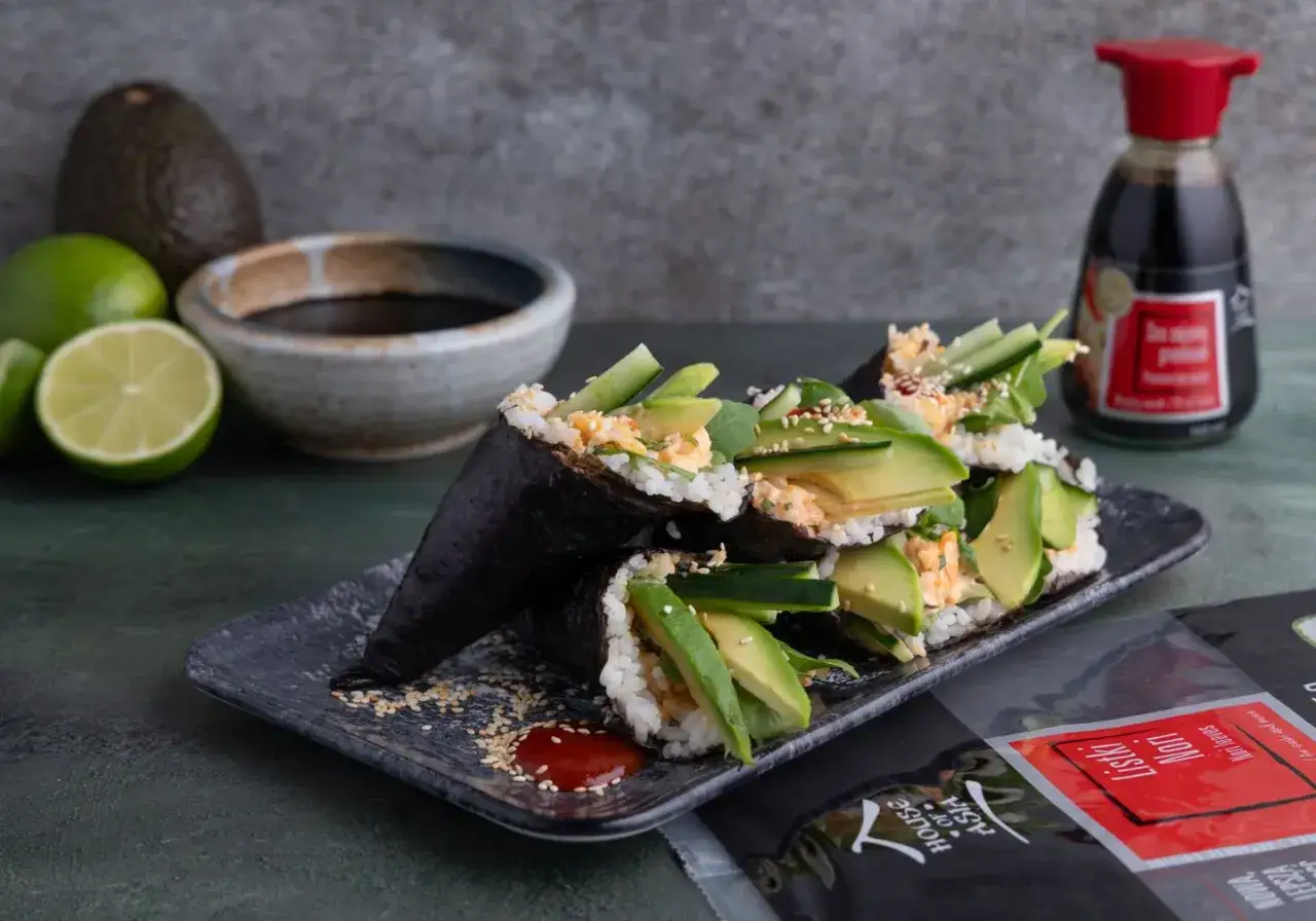 Smaczne temaki sushi z awokado, og&oacute;rkiem i pikantnym farszem, podane na czarnym talerzu z sosem sojowym i limonkami w tle.