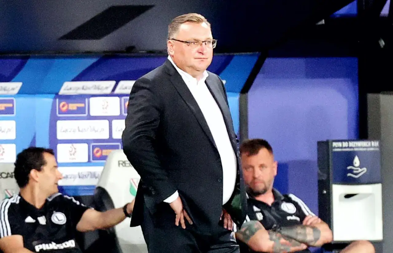 Polski trener w garniturze stoi na tle ławki rezerwowych. Obok siedzą dwaj mężczyźni w strojach sportowych.