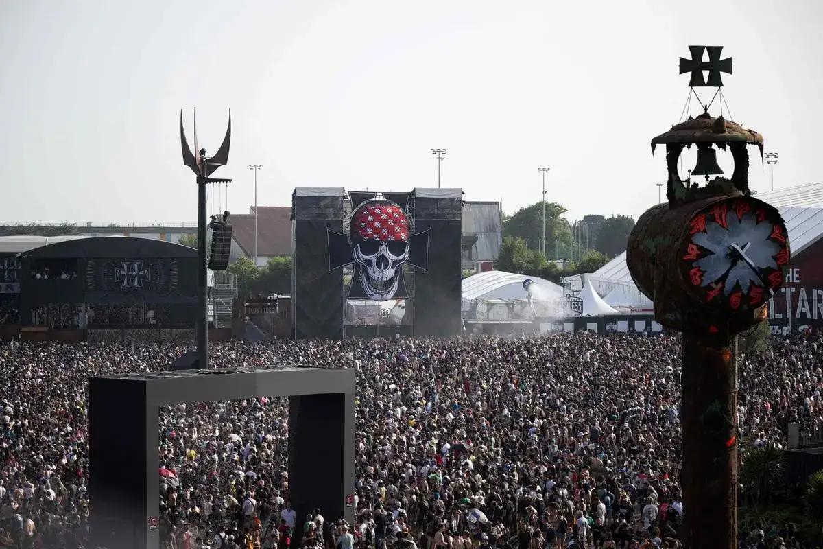 Foule immense devant une scène de festival de musique metal. Un crâne bandeau rouge domine l'arrière-plan. Un groupe metal actuel en pleine action.