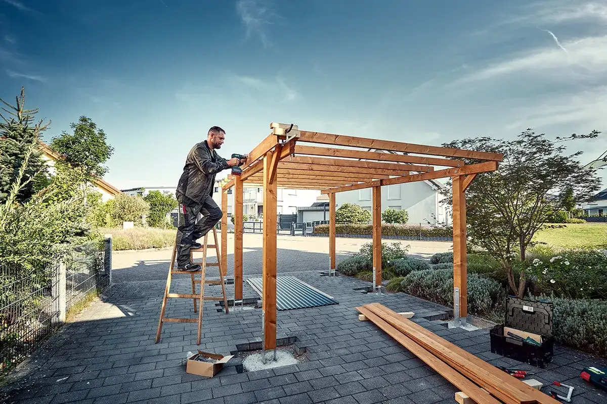 Man bygger en carport på plintar. Han står på en stege och skruvar fast takbjälkar.