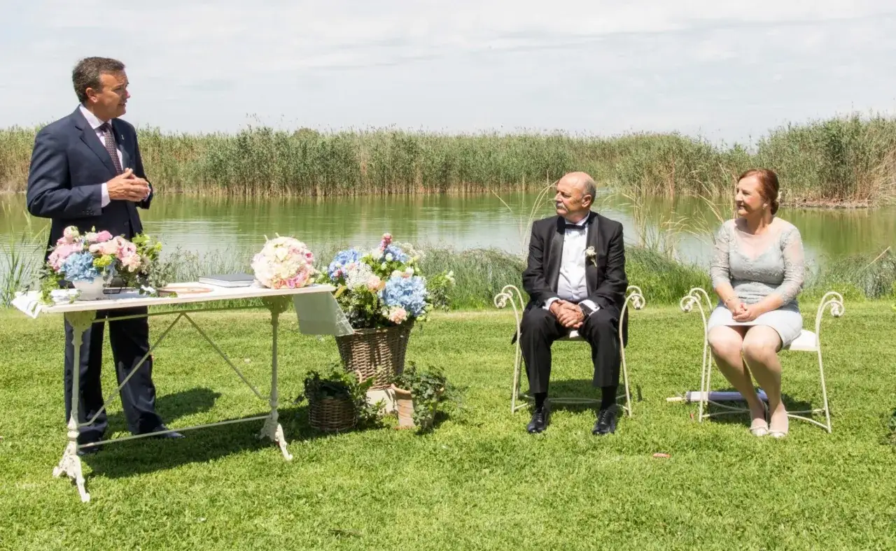 Ceremonia al aire libre junto a un lago. Un hombre habla, mientras una pareja sentada espera. Ideal para frases para bodas de oro cortas.