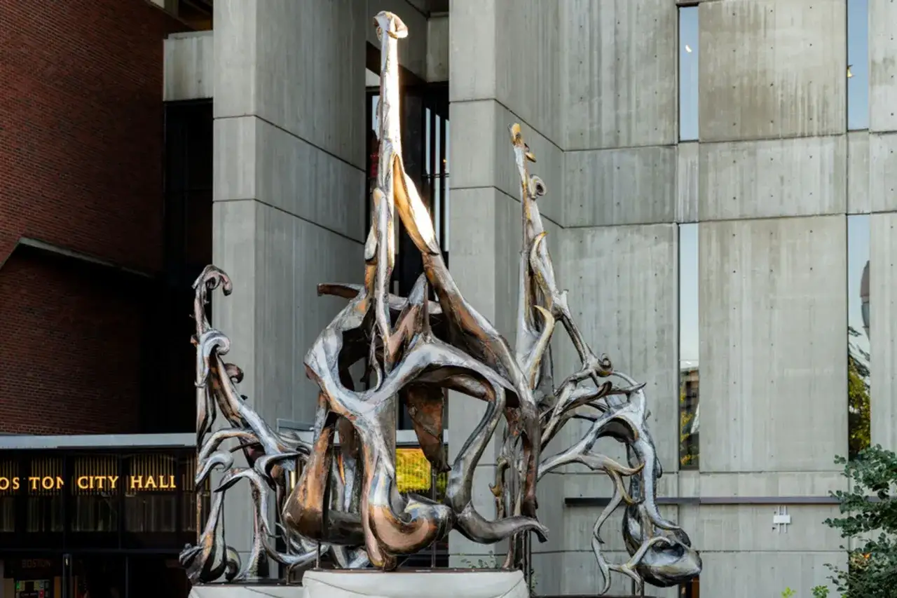 A dynamic, abstract metal sculpture stands as a public art installation outside Boston City Hall, its organic forms catching the light.