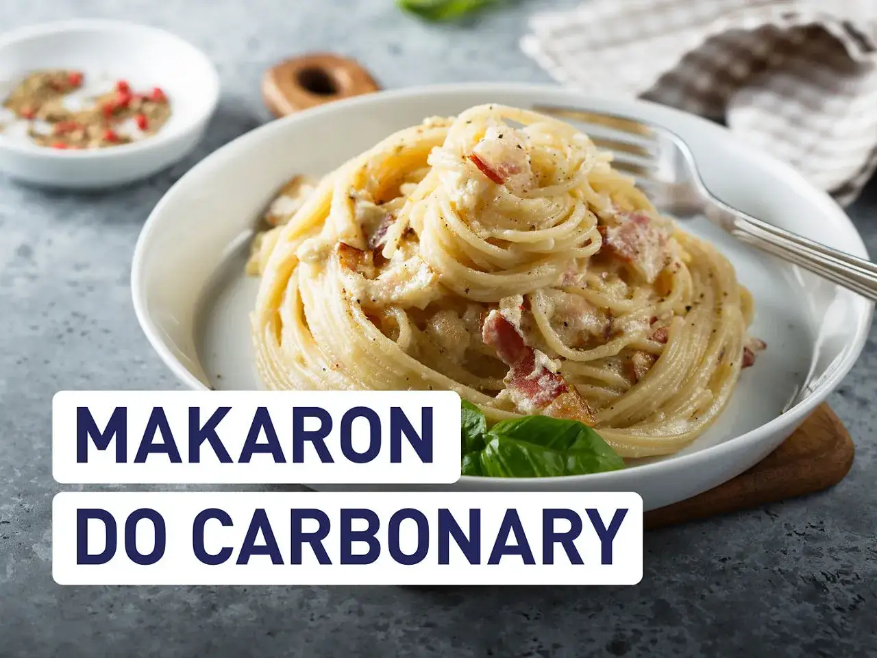 Pyszny makaron do carbonary z boczkiem i kremowym sosem, podany w białej misce. Obok przyprawy i liść bazylii.