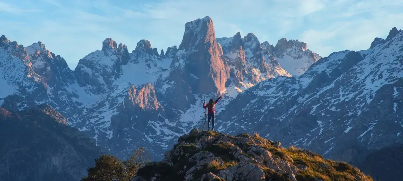 Una excursionista celebra en un mirador de los Picos de Europa, con picos nevados iluminados por el sol poniente.