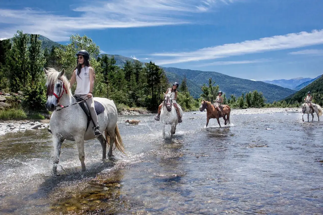Cuatro jinetes disfrutan de rutas en caballo por un río cristalino, rodeados de montañas y bosques frondosos.